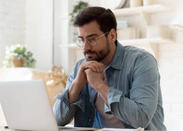 Pensive young Caucasian man in glasses sit at desk look at laptop screen thinking pondering, thoughtful millennial male in spectacles work at computer consider ideas solving problems at home workplace